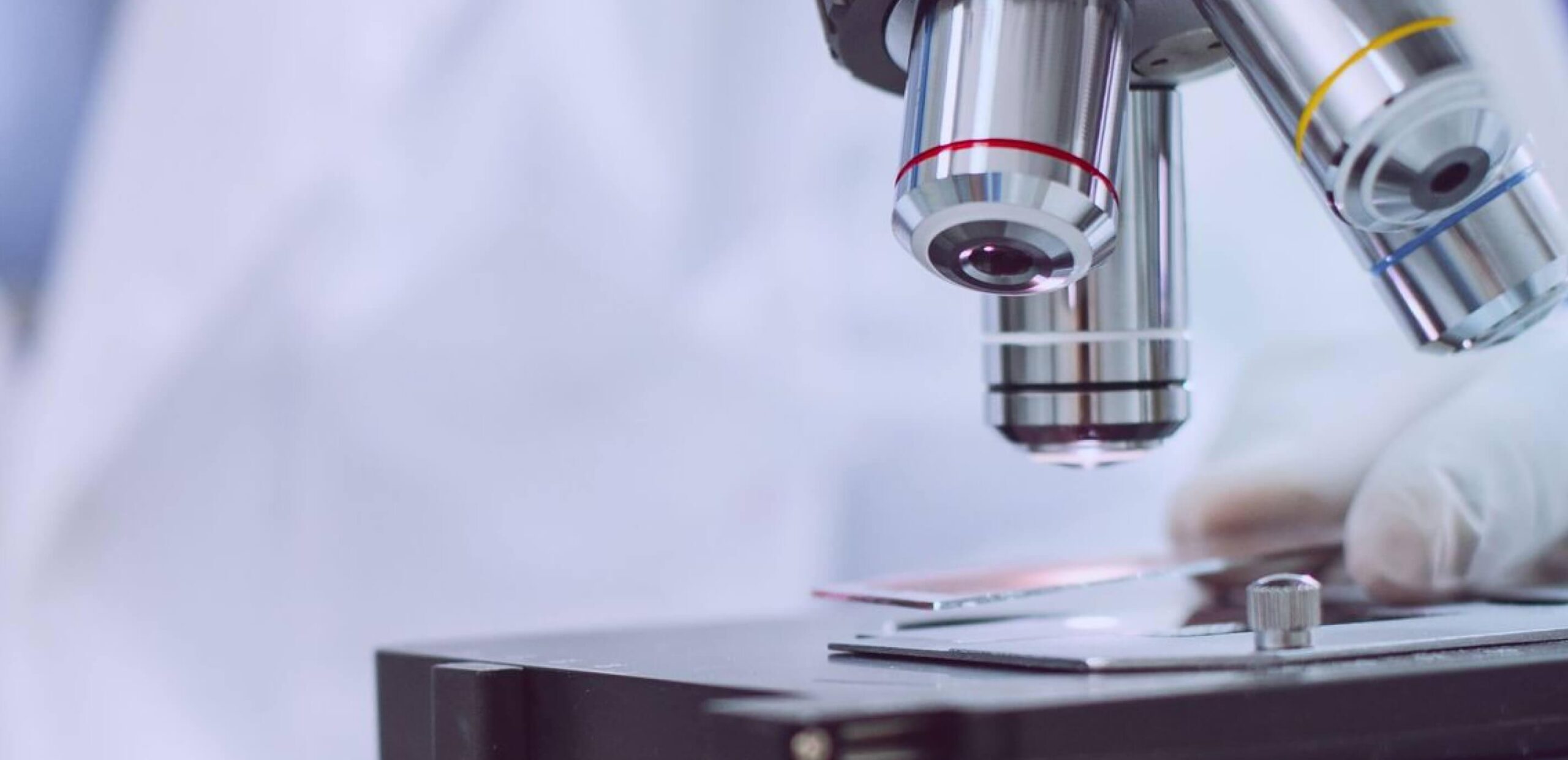 Close-up of microscope objective lenses focused on a glass slide, with a gloved hand adjusting the specimen on the stage.