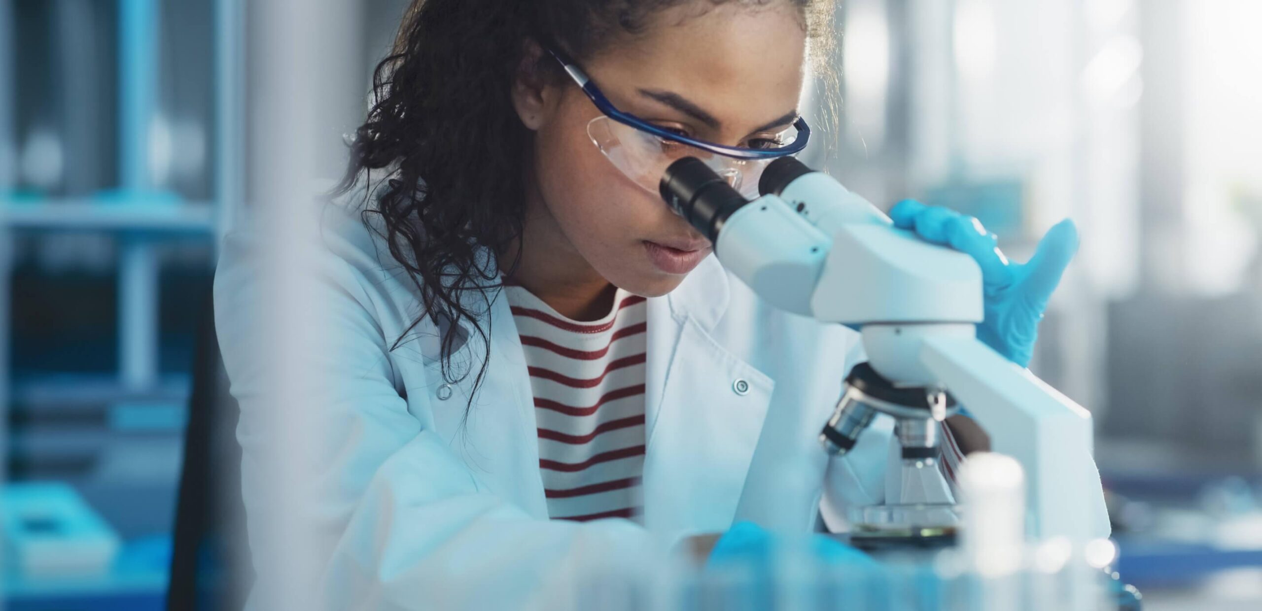 Scientist wearing protective goggles and gloves looking into a microscope in a laboratory environment.