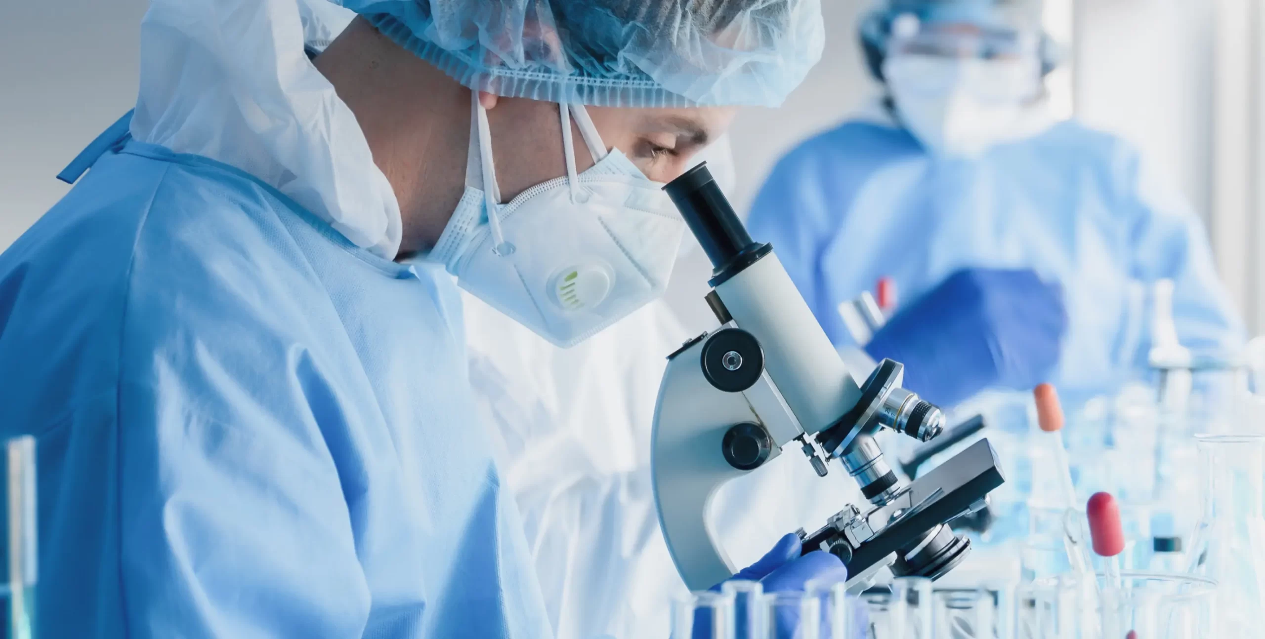 Scientist in protective clothing and face mask using a microscope in a laboratory, with lab glassware and a colleague in the background.