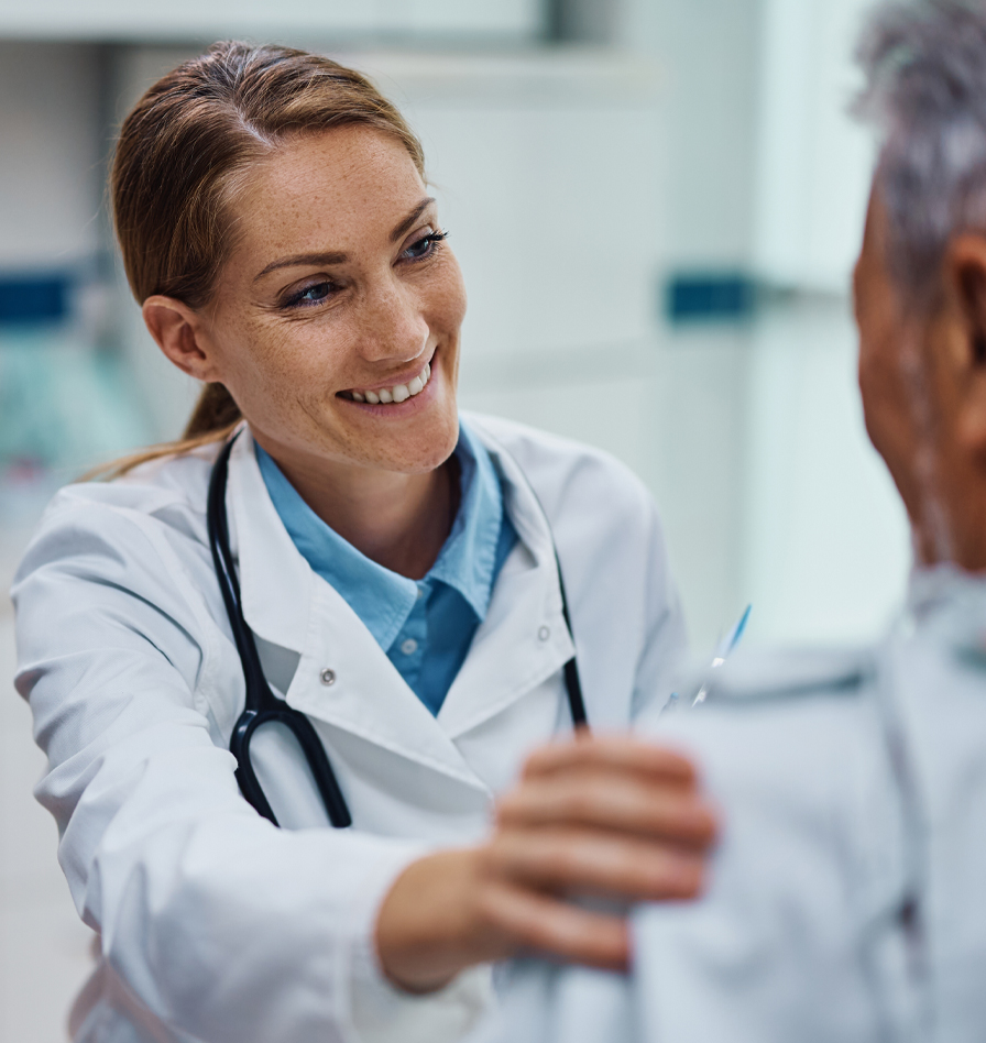Female doctor wearing a white lab coat and stethoscope smiling and placing a hand on a patient's shoulder. The patient is partially visible from behind, and the interaction takes place in a bright medical setting.