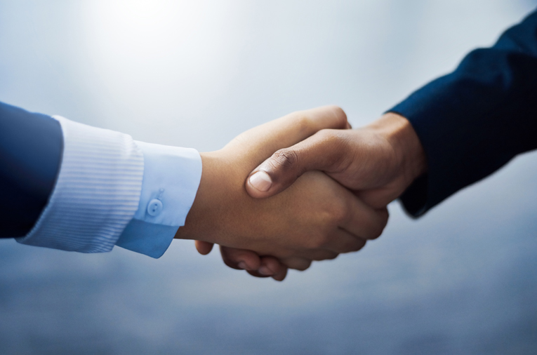 Close-up of two people shaking hands, one wearing a dark suit and the other a light blue shirt with a white cuff. The background is softly blurred with a cool blue tone.