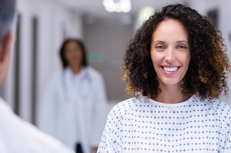 Woman with shoulder-length curly hair wearing a hospital gown stands in a hallway, smiling at the camera. A person in a white coat is blurred in the background, and another individual is partially visible in the foreground, seen from behind.