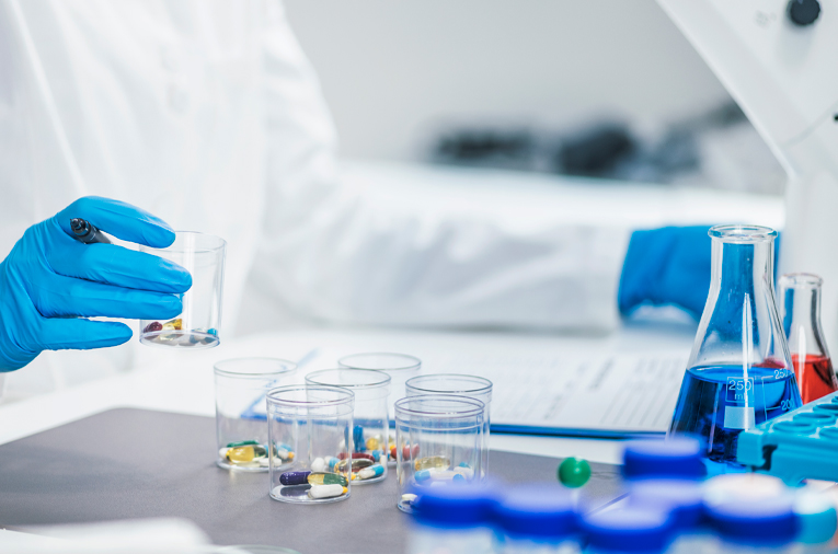Laboratory setting with a person in a white lab coat and blue gloves holding a clear plastic container filled with capsules and tablets. Several similar containers with assorted pills are arranged on the table. Also visible are a blue liquid-filled flask, a red solution in a conical flask, and laboratory documents on a clipboard.