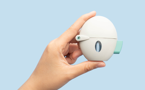 Hand holding a compact, oval-shaped medical inhaler with a small transparent window and a green lever, set against a light blue background.