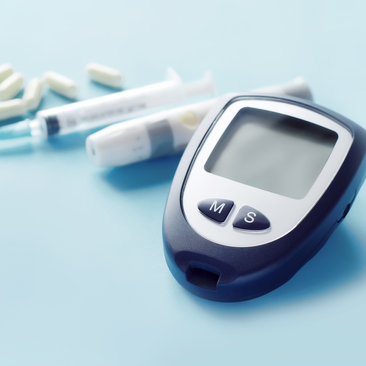 Close-up of a digital blood glucose meter with two buttons labeled 'M' and 'S' on a light blue surface. In the blurred background are a syringe, an insulin pen, and several white capsules.