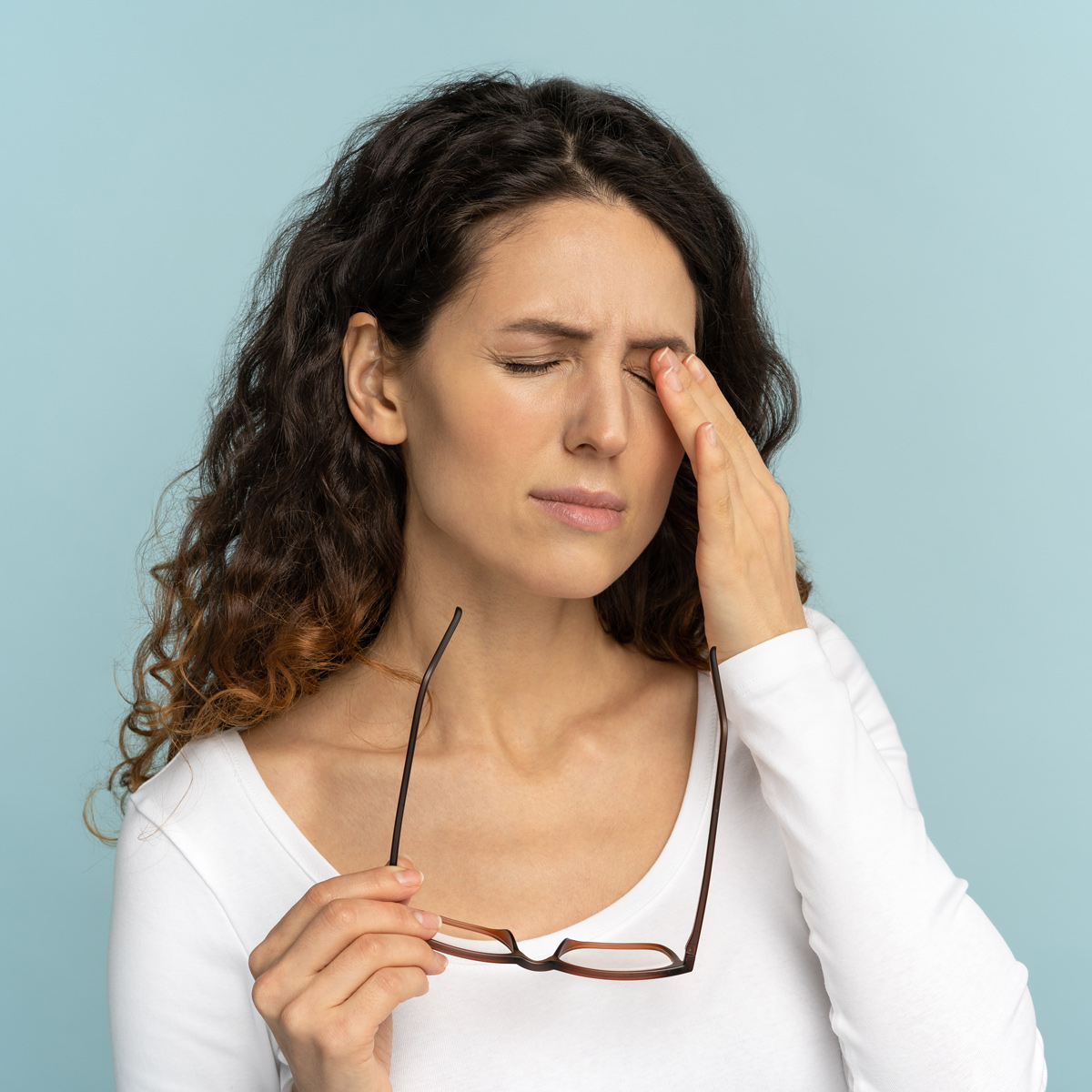Woman with long curly brown hair wearing a white shirt, holding eyeglasses in one hand and touching her closed right eye with the other, against a light blue background.