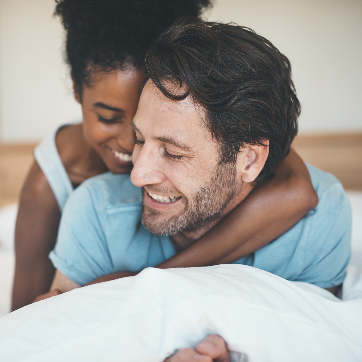 A woman with curly hair tied up is hugging a man with medium-length brown hair and a beard from behind. Both are smiling and wearing light-colored clothing while lying on a white bed.