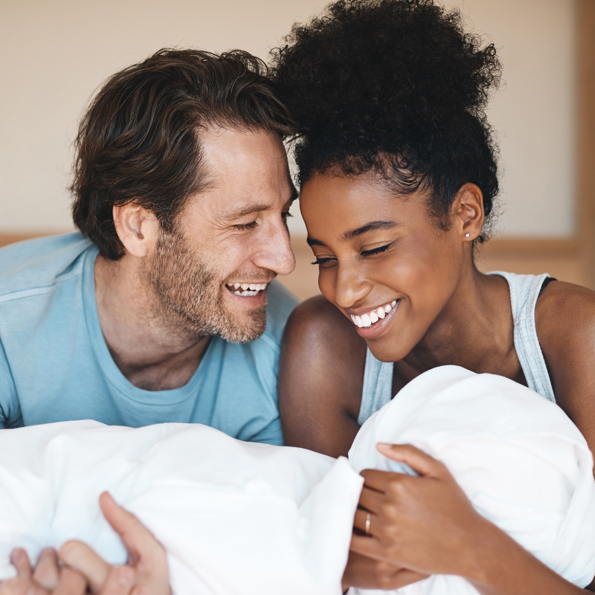 A man with medium-length brown hair and a beard, and a woman with curly hair tied up, are looking down at an infant wrapped in a white blanket. The man wears a light blue shirt, and the woman wears a sleeveless gray top.