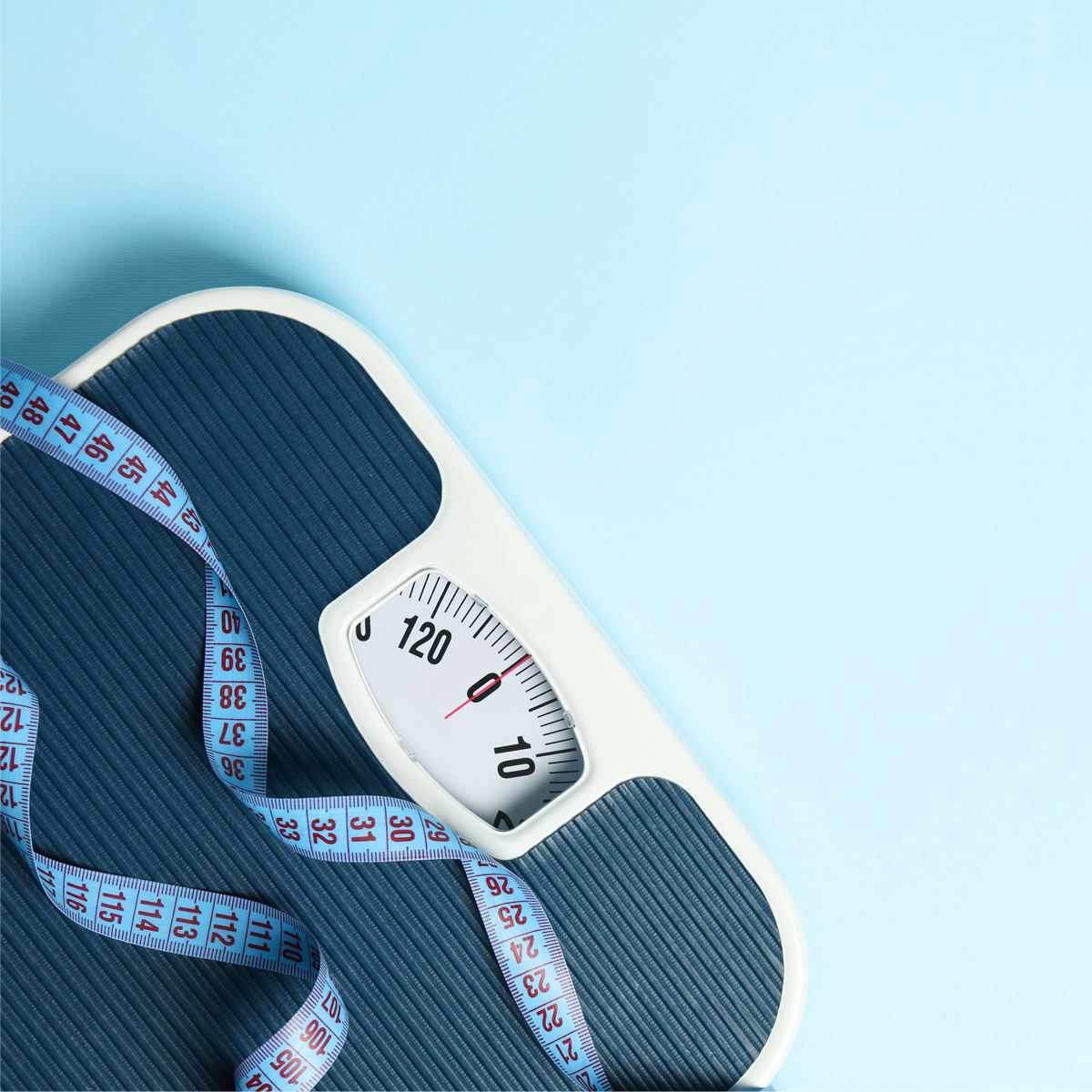 Partial top-down view of a mechanical bathroom scale with a white border and dark blue textured surface, displaying a zero reading in kilograms. A blue measuring tape with red numbers is loosely draped across the scale. The background is light blue.