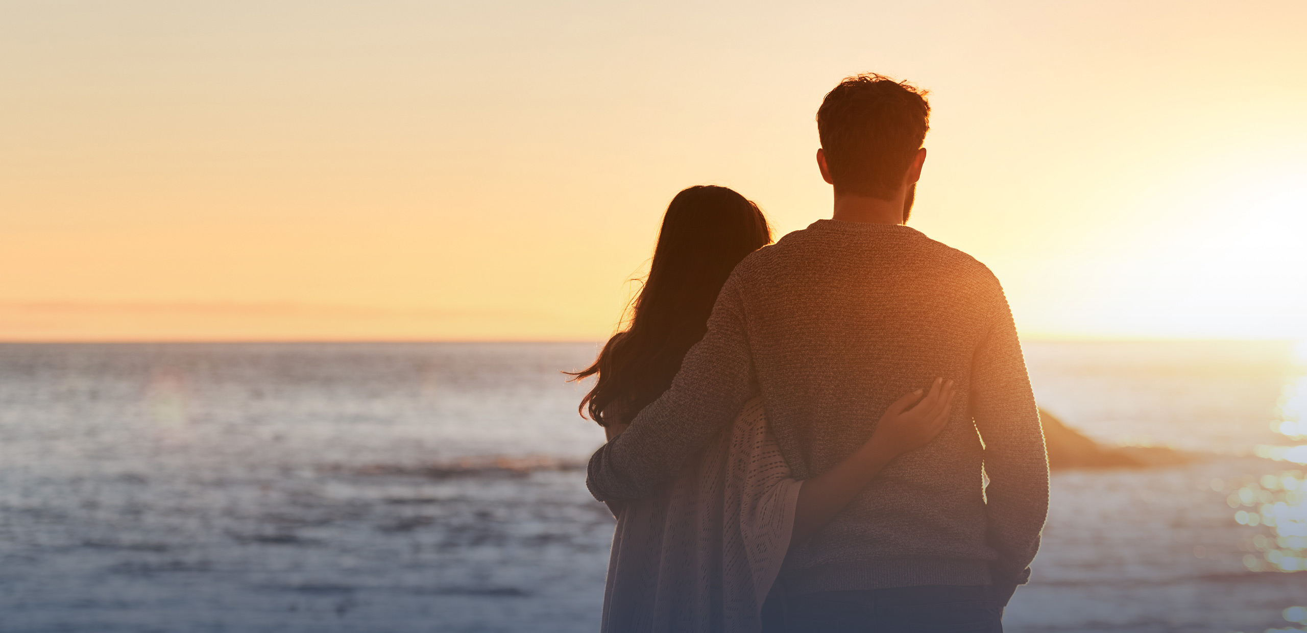Man and woman standing with arms around each other, looking at the ocean during sunset.