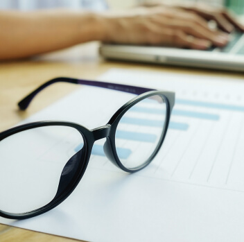 Black eyeglasses resting on a printed bar chart, with a person typing on a laptop in the blurred background.