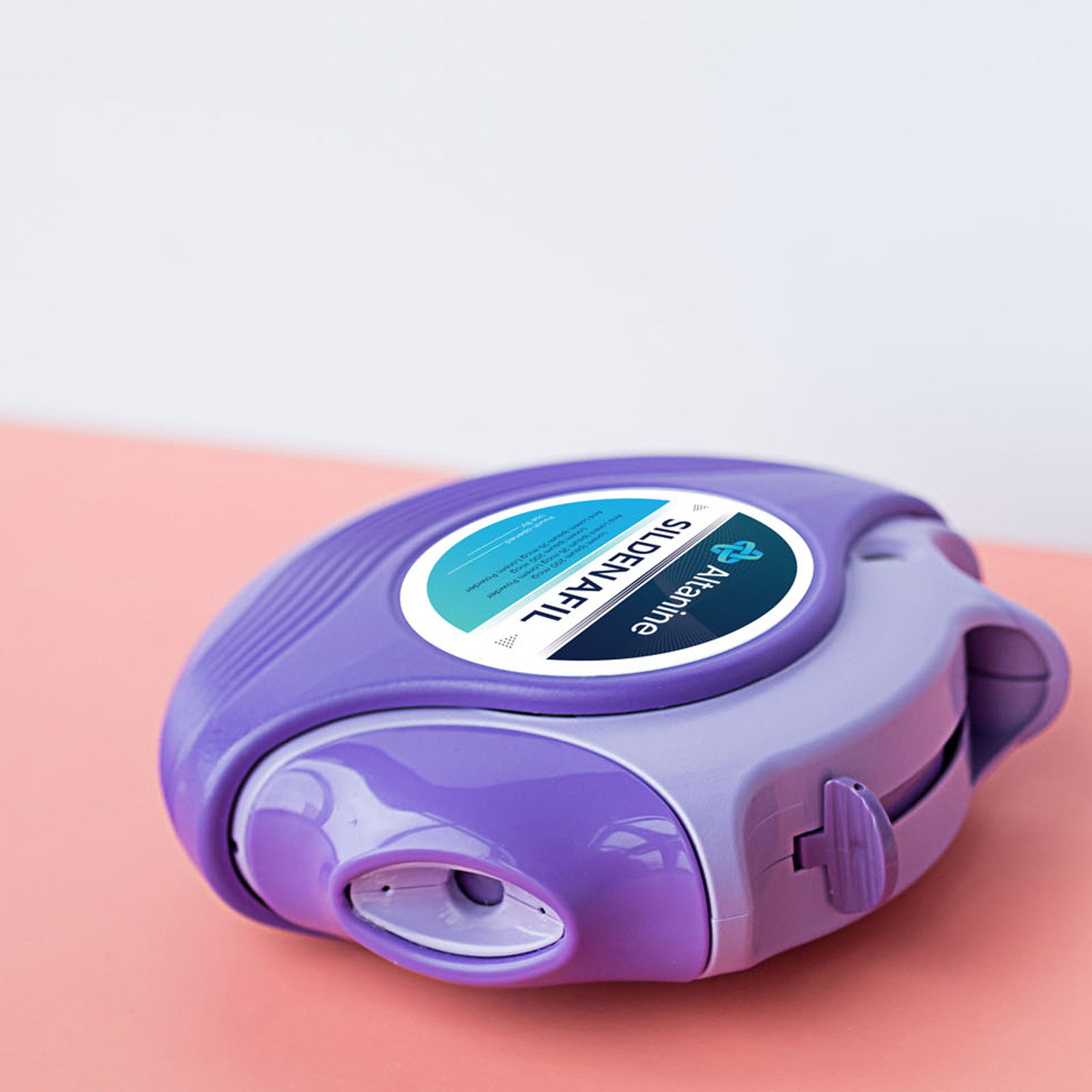 A close-up photo of a purple and lavender circular plastic medical inhaler device with a white and blue label on top. The label reads 'Altanine' and 'SIDENAFIL' in bold text, indicating the brand and medication name. The inhaler rests on a smooth coral-pink surface with a plain white background.