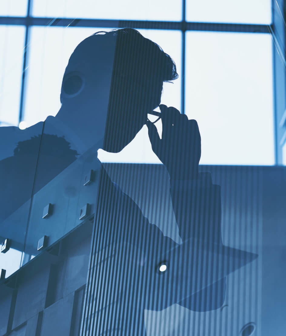 Silhouette of a person holding glasses near their face, seen through a reflective glass surface with architectural and window reflections in blue tones.
