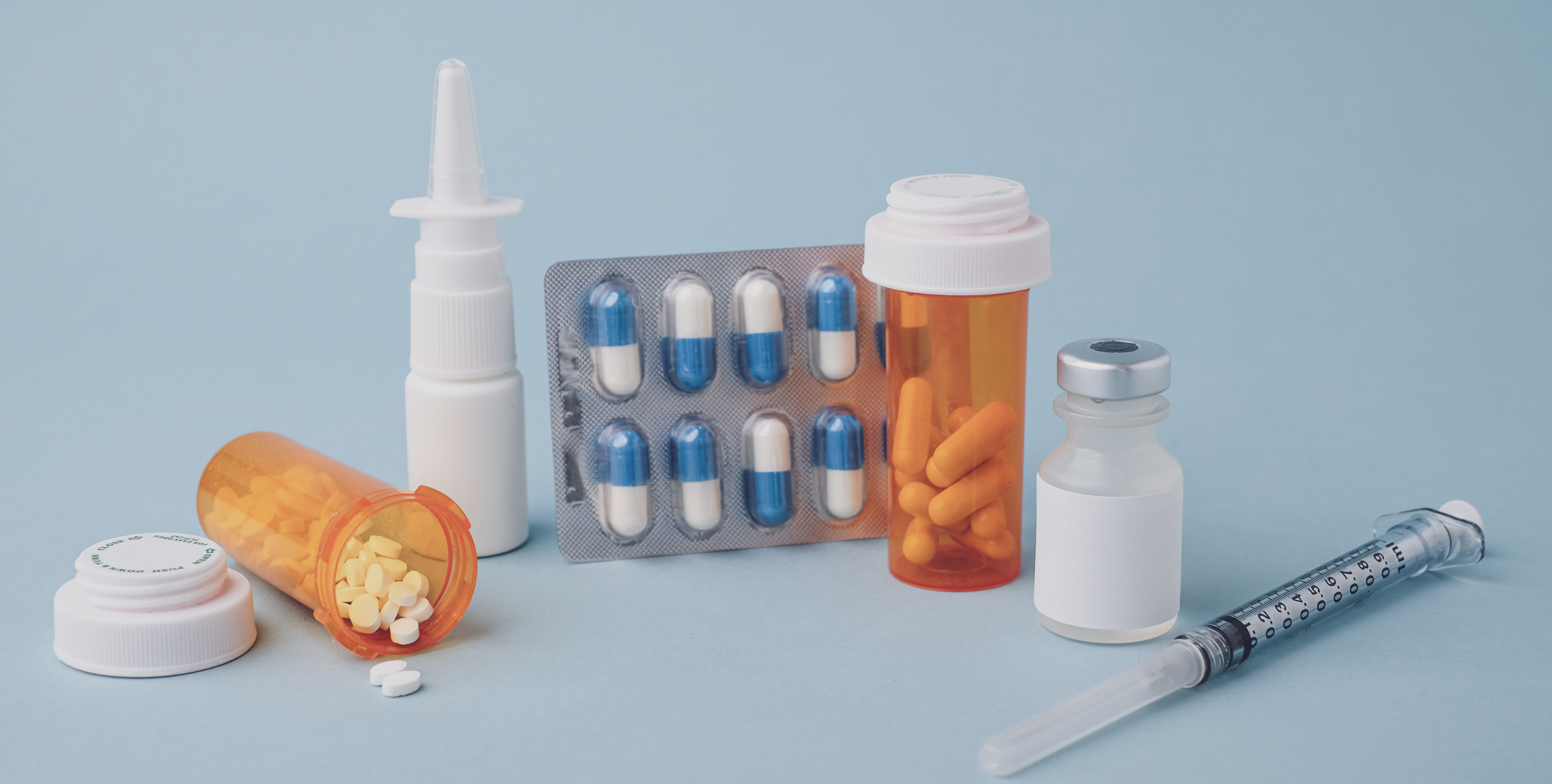 Assorted pharmaceutical items on a light blue background, including orange pill bottles with tablets and capsules, a blister pack of blue-and-white capsules, a nasal spray, a vaccine vial, and a syringe, representing various forms of medication delivery.