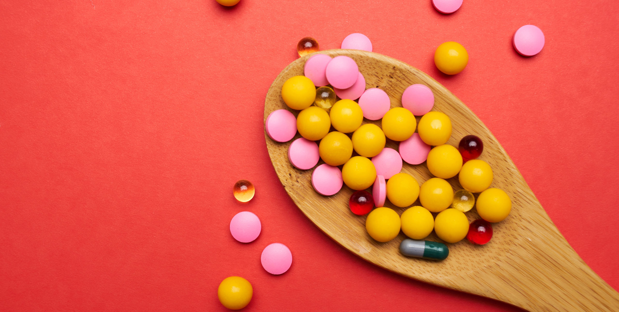 Assorted colorful pills and capsules, including yellow, pink, red, and gel tablets, displayed on a wooden spoon against a vibrant red background, symbolizing variety in pharmaceutical or nutritional supplements.