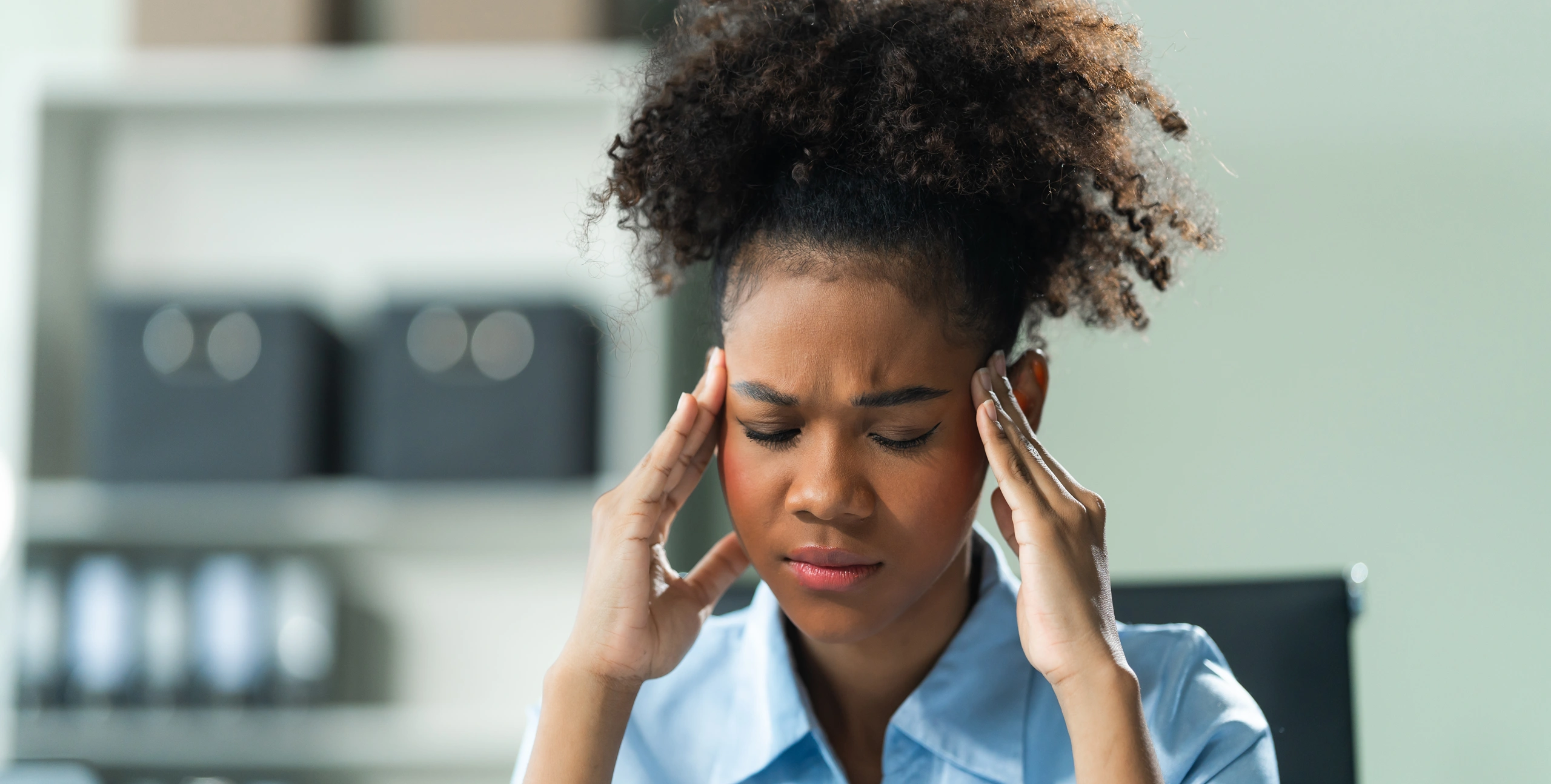 Woman with curly hair touching her temples with both hands, eyes closed and brow furrowed, showing visible discomfort from a headache or migraine. She is seated indoors with shelves and office items blurred in the background.