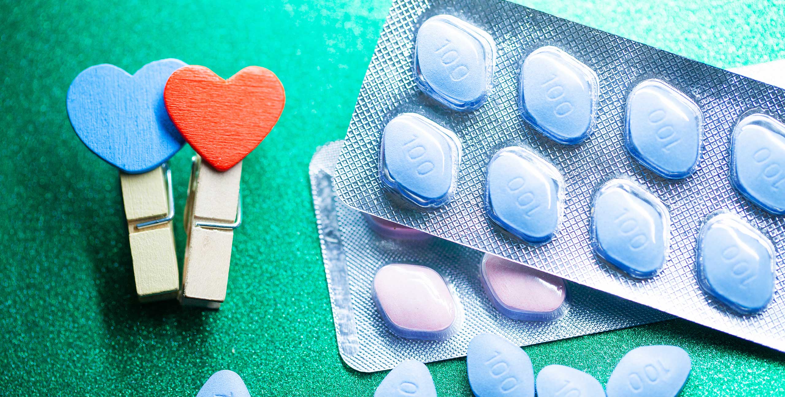 Blister packs of blue tablets marked ‘100’ arranged on a shimmering green surface, next to two wooden clothespins topped with blue and red heart shapes, symbolizing love and medication—possibly representing treatments for erectile dysfunction or cardiovascular health.
