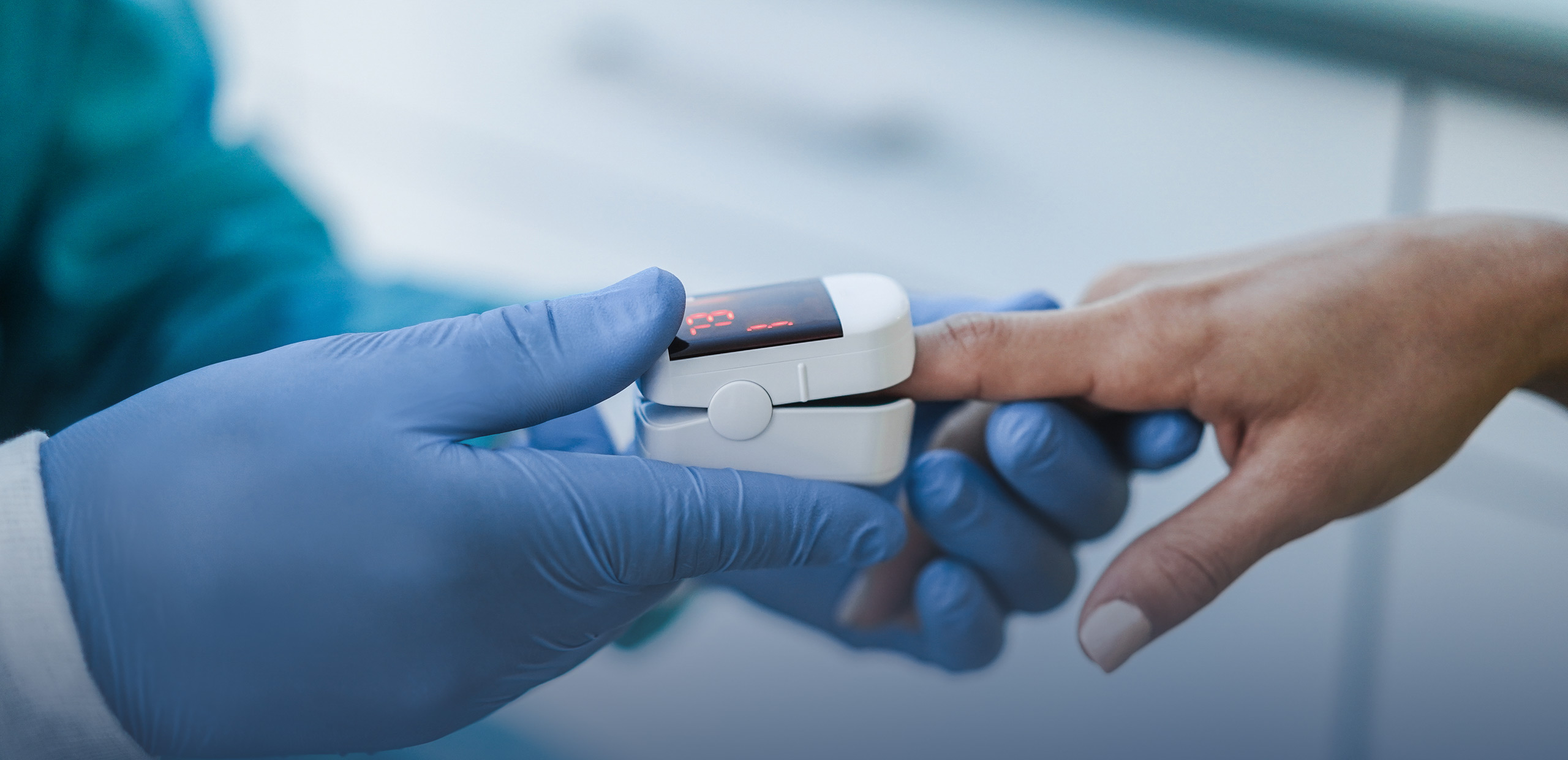 Healthcare professional wearing blue gloves placing a fingertip pulse oximeter on a patient’s finger to measure oxygen saturation and heart rate, in a clinical environment.