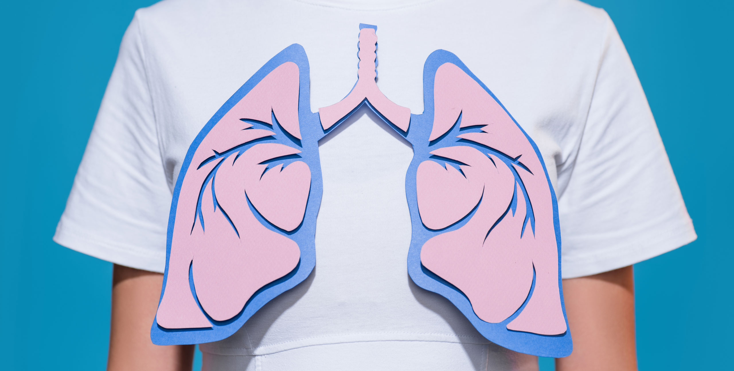 Person wearing a white shirt with a stylized paper-cut illustration of pink and blue lungs placed over the chest, representing respiratory health, against a bright blue background.