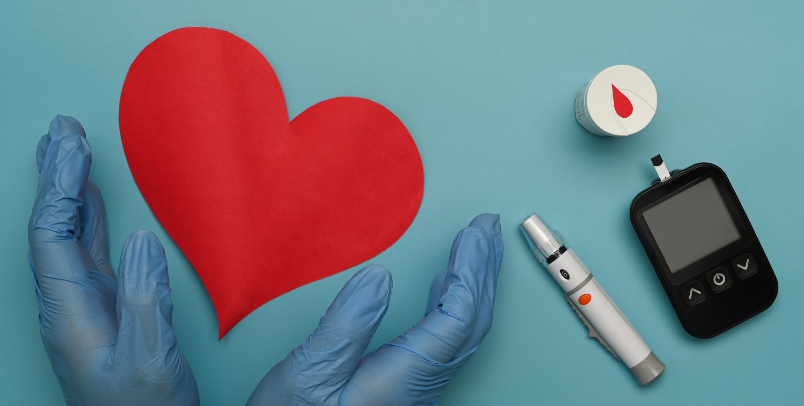 Blue-gloved hands surrounding a red paper heart, with a blood glucose meter, test strip container, and insulin pen on a blue background, symbolizing diabetes care and heart health.