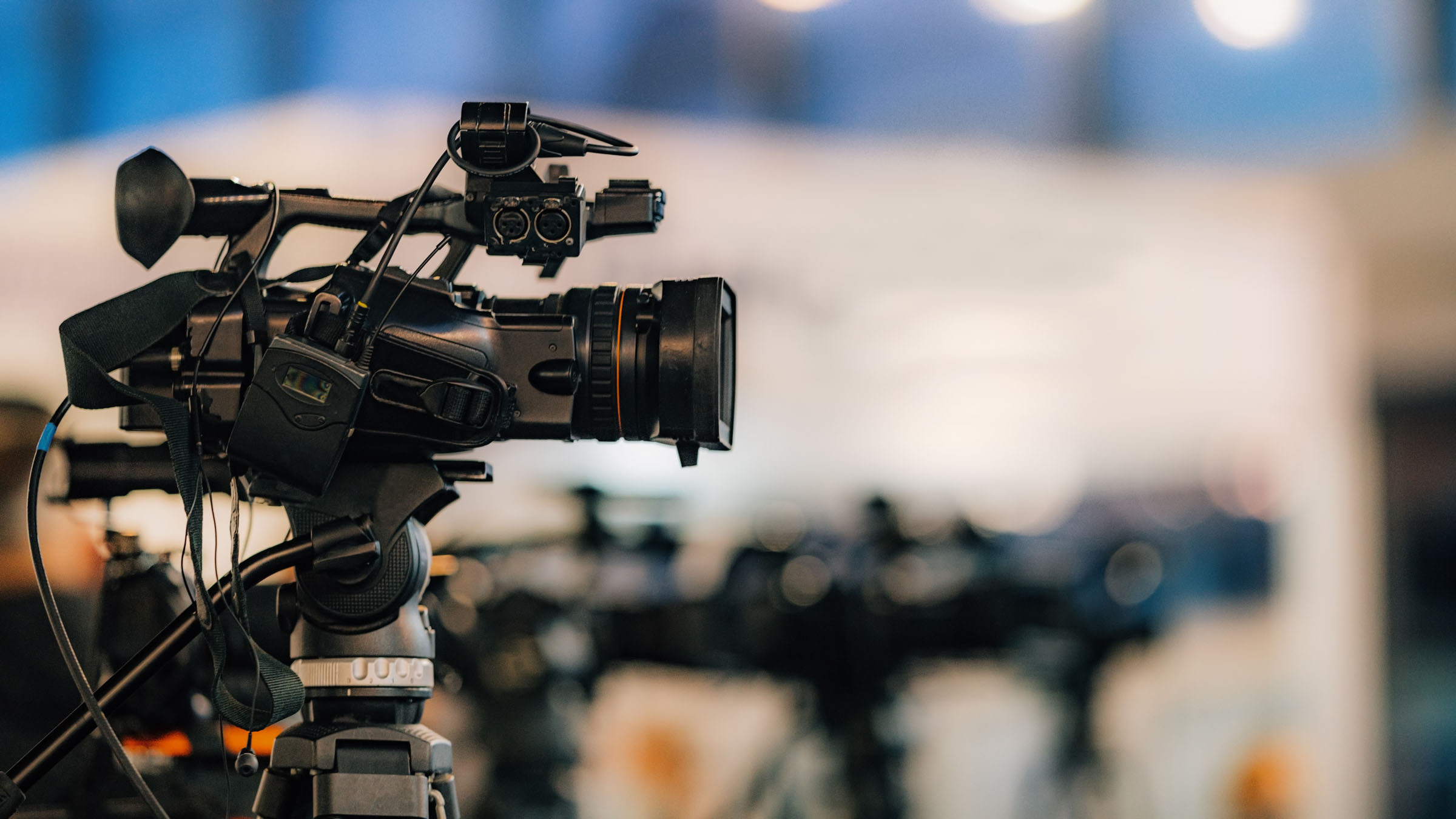 Professional video camera on tripod at media event or press conference with blurred background of broadcast equipment.