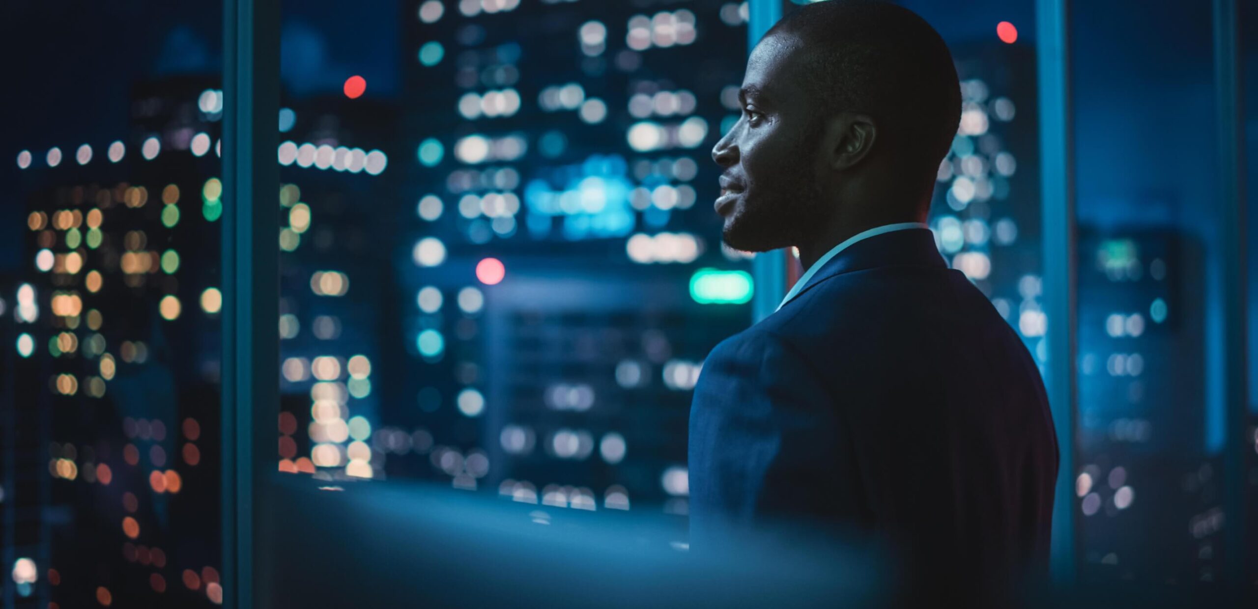 Man in a dark suit looking out of a window at night with blurred city lights in the background.
